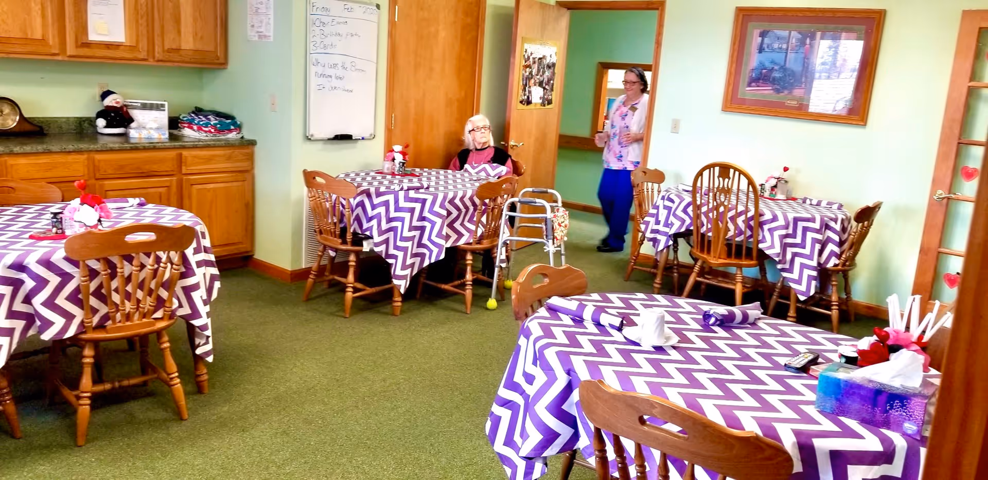 A dining room with several wooden tables covered with purple and white chevron-patterned tablecloths. Wooden chairs surround the tables. An elderly woman sits at one table with a walker nearby, and another person is walking through a doorway in the background. The room has light green walls, wooden cabinets, and a whiteboard on one wall.