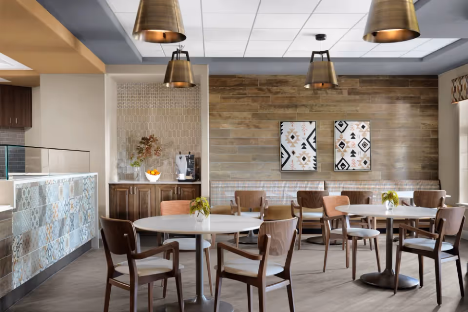 A modern dining area with round white tables and wooden chairs. The back wall features wood paneling and two framed abstract artworks. There are three brass pendant lights hanging from the ceiling. A small counter area with patterned tiles and a coffee machine is visible on the left side. The room has a warm and inviting atmosphere with natural light coming from a window on the right.