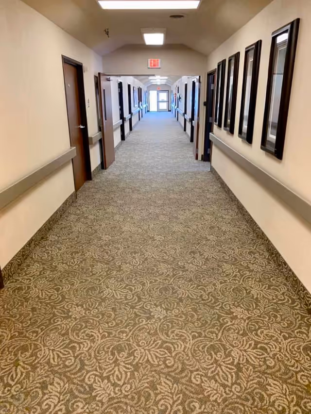 Long carpeted interior hallway with doors on both sides, handrails along the walls, and framed artwork on the right.