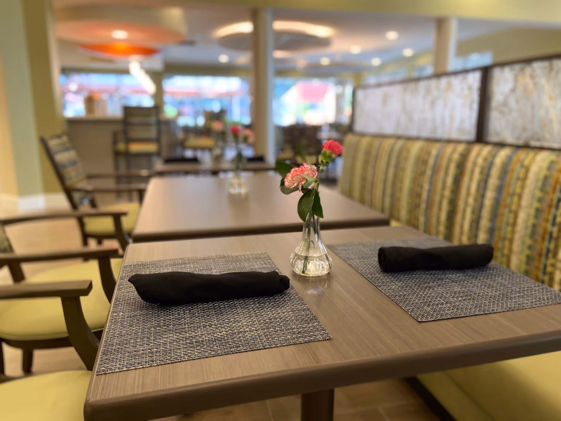 A dining area with tables set with black rolled napkins on woven placemats and small glass vases holding pink flowers. The seating includes cushioned chairs and a striped upholstered bench. The background shows more tables and chairs with soft lighting.