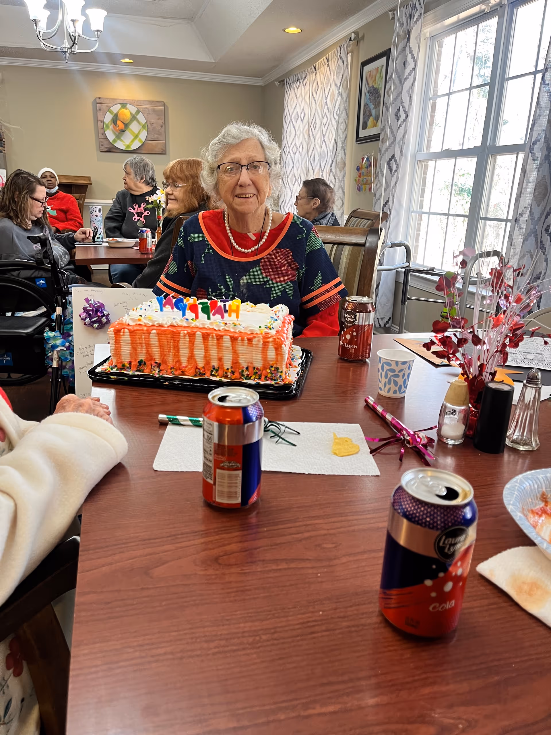 An elderly woman smiles behind a decorated birthday cake at a communal dining table with other residents.