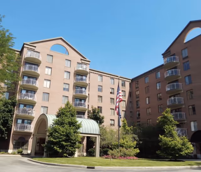 Exterior view of a multi-story senior living facility building with balconies, a covered entrance, and landscaped greenery including trees and bushes. Two flagpoles with American and state flags are visible in front of the building under a clear blue sky.