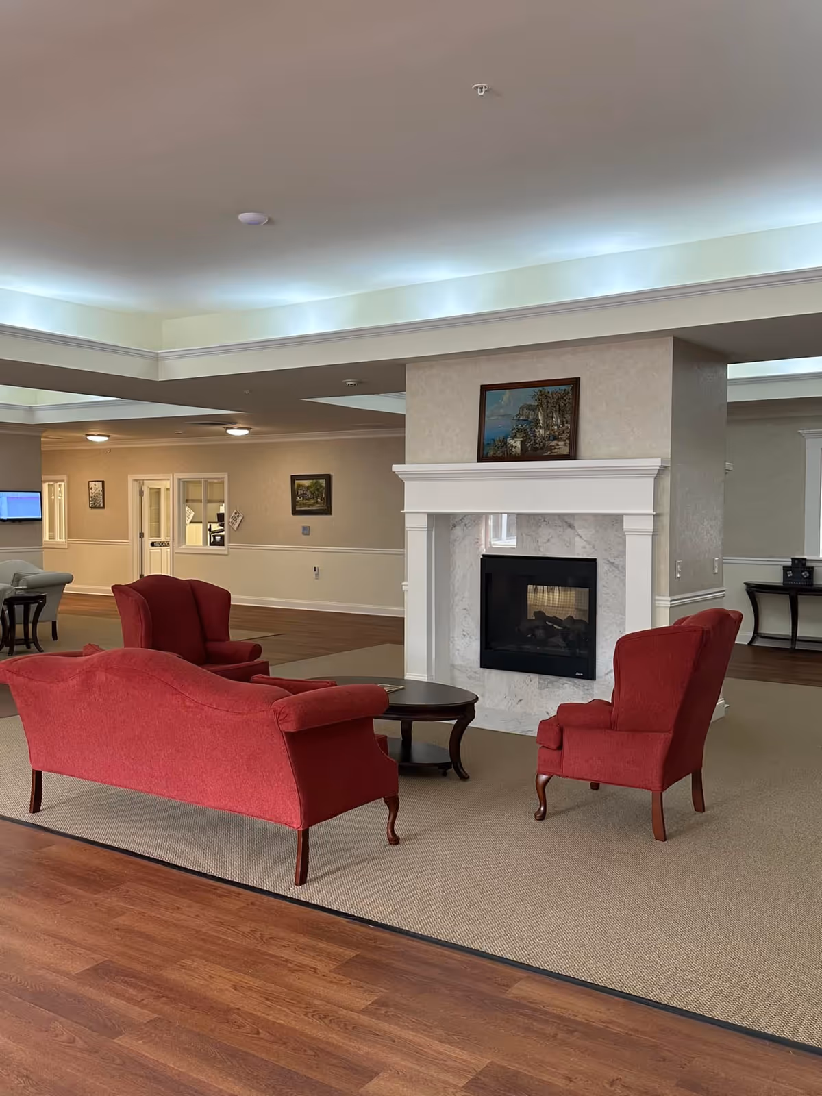 A cozy seating area in a senior living facility with a red sofa and two red armchairs arranged around a dark wooden coffee table in front of a white fireplace with a marble surround. A framed painting hangs above the fireplace. The room has beige walls, wood flooring, and recessed lighting in the ceiling.