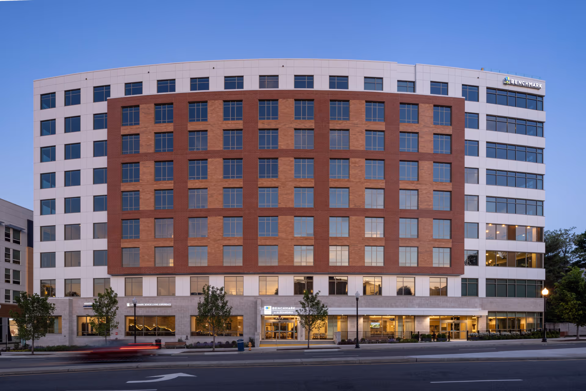 Exterior view of a multi-story senior living facility building named Benchmark at Alexandria during early evening with clear sky, showing large windows and entrance area with trees and street in front.