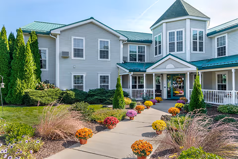Exterior view of a two-story assisted living facility with light gray siding and green roofs. A concrete walkway lined with colorful potted flowers leads to the main entrance, which has a covered porch. The landscaping includes various shrubs, small trees, and ornamental grasses under a clear blue sky.