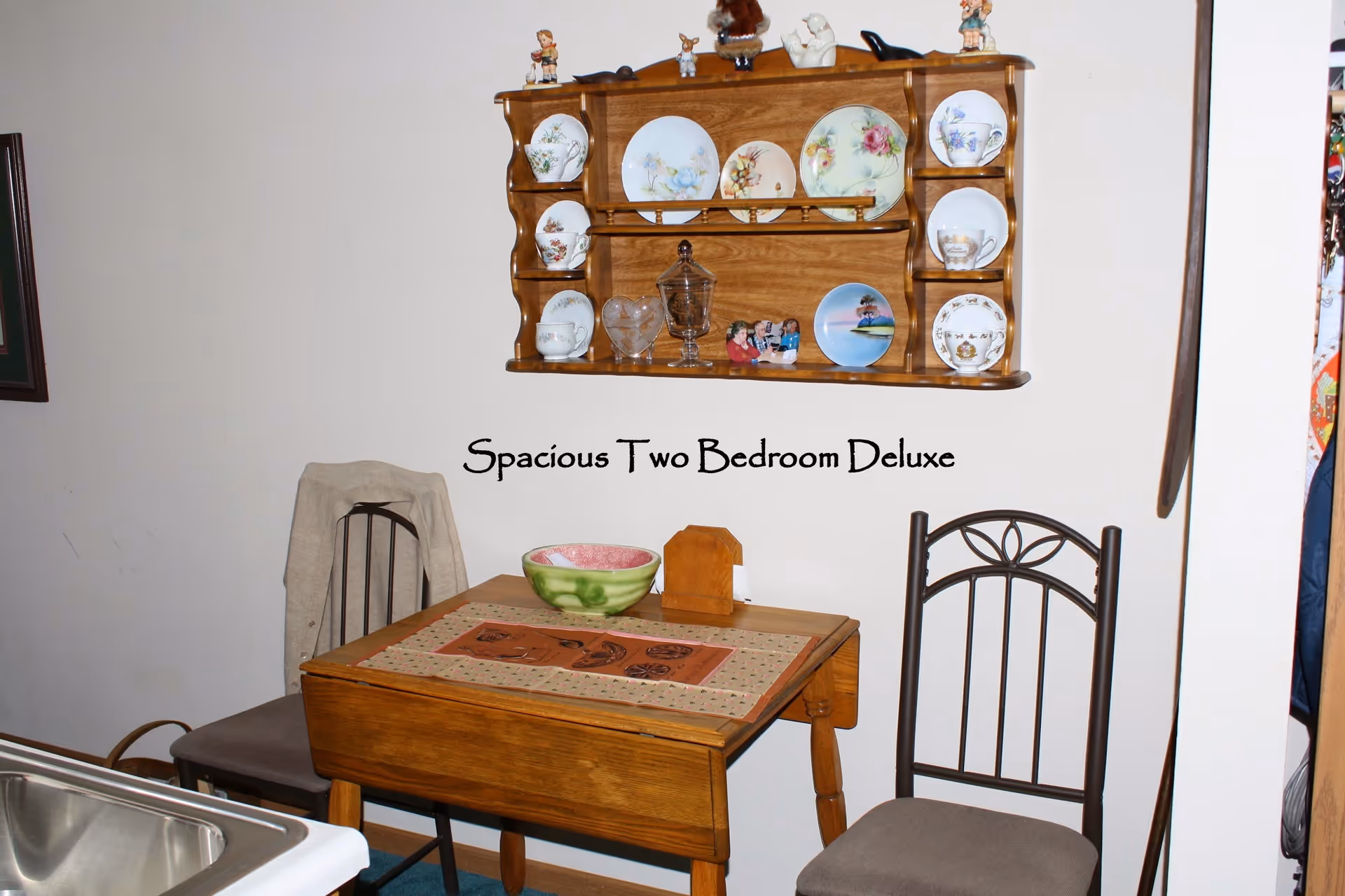 Small dining area with a wooden table and two chairs beneath a wall-mounted plate rack and text reading 'Spacious Two Bedroom Deluxe'.