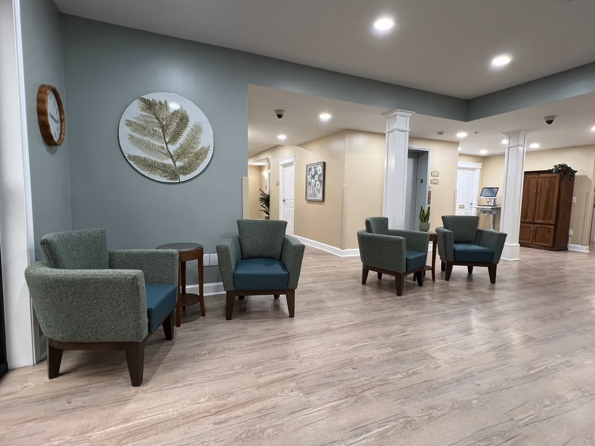 Spacious senior living common area with several green upholstered armchairs, small tables, decorative wall art, and wood-look flooring.