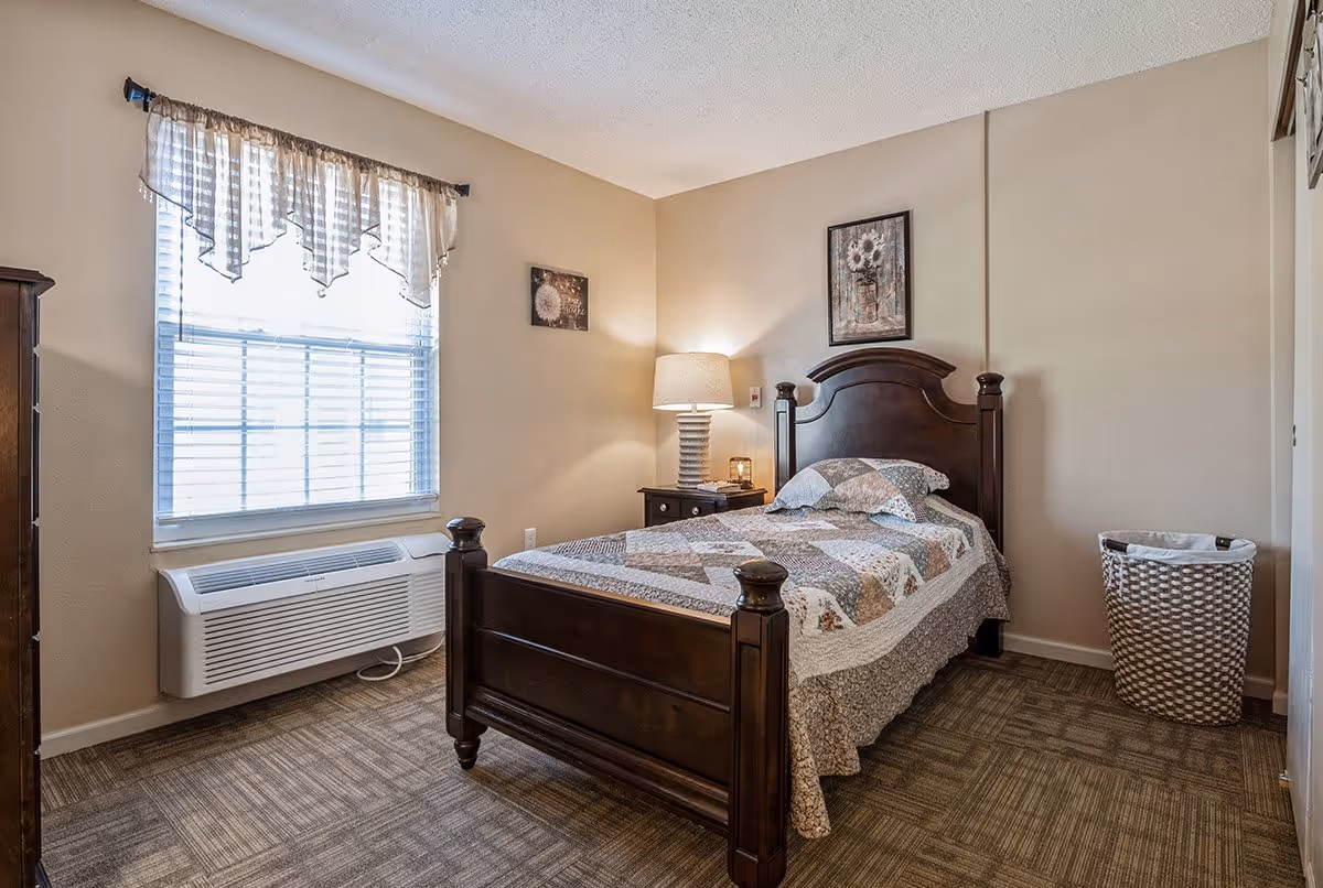 A cozy bedroom with a single dark wooden bed frame, a patchwork quilt, and a matching pillow. There is a nightstand with a lamp and a small decorative item next to the bed. A window with blinds and a valance curtain lets in natural light. The room has beige walls, carpeted flooring, a framed floral picture above the bed, and a woven laundry basket in the corner.