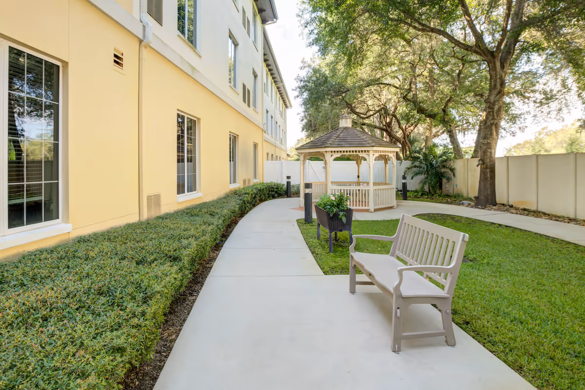 Outdoor garden area at Arbor Terrace Citrus Park featuring a paved walkway, a beige bench, a planter with flowers, a white gazebo, green grass, and large trees providing shade, next to a yellow building with multiple windows.