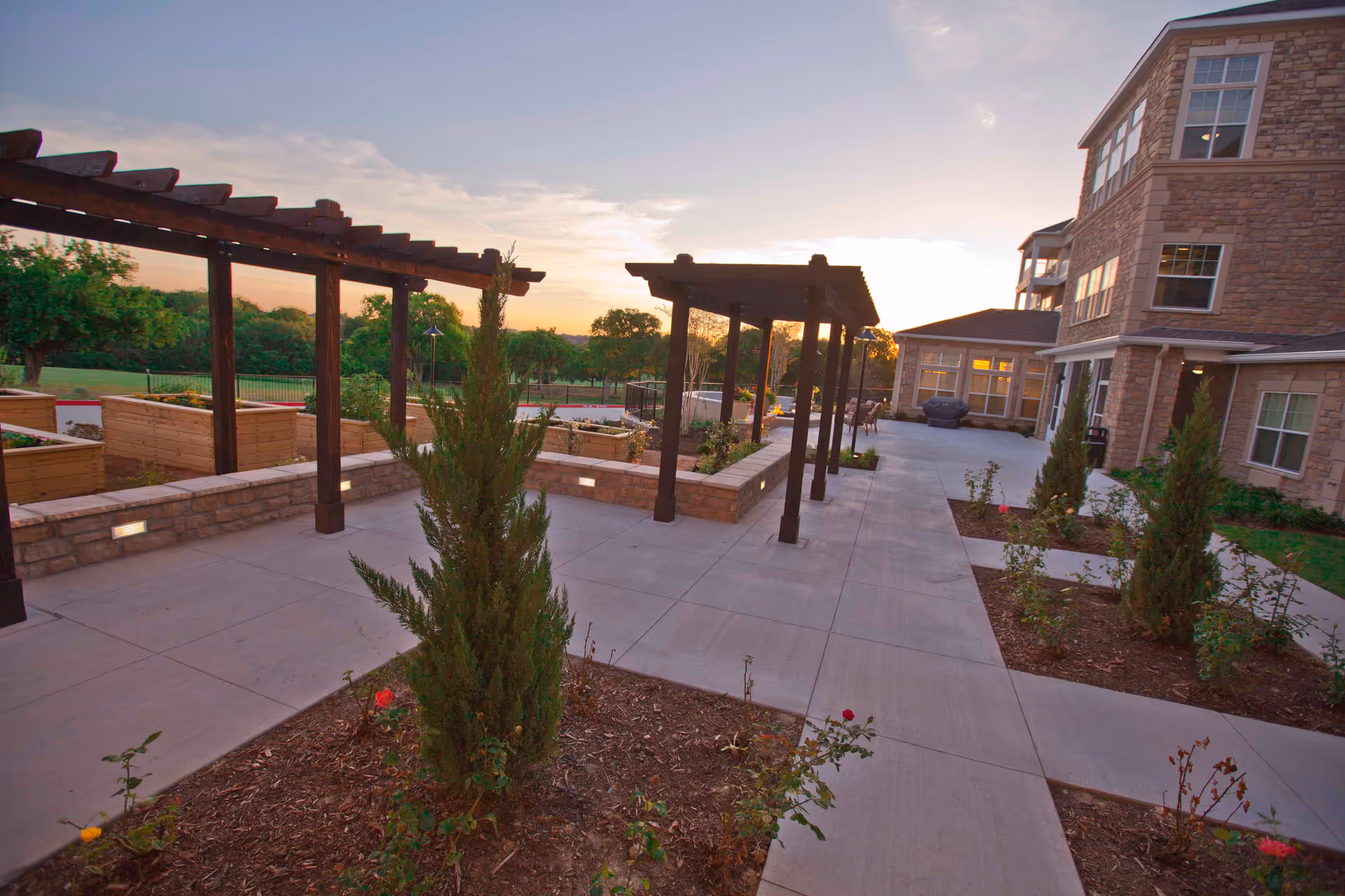 Outdoor patio area at Pioneer Ridge Gracious Retirement Living featuring wooden pergolas, raised garden beds, small trees, and shrubs with a stone building in the background during sunset.