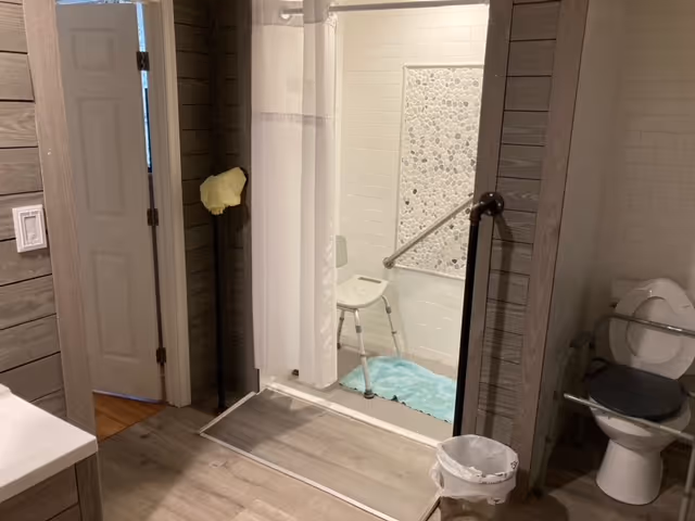 Accessible bathroom with a roll-in shower, shower chair, grab bar, and a toilet with a raised seat.