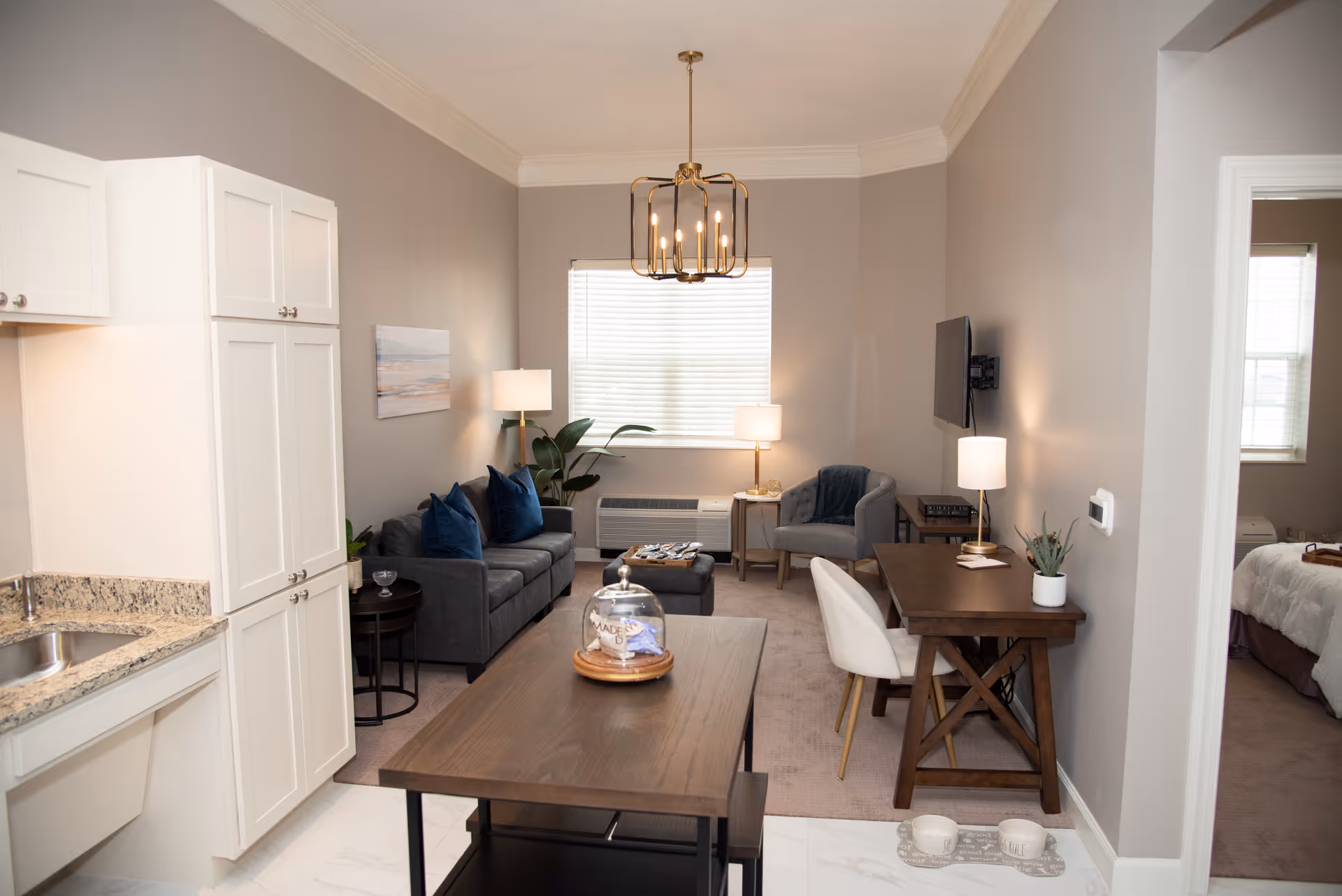 Bright open-concept living room with a gray sofa, armchair, wall-mounted TV, desk, and a kitchenette visible on the left.