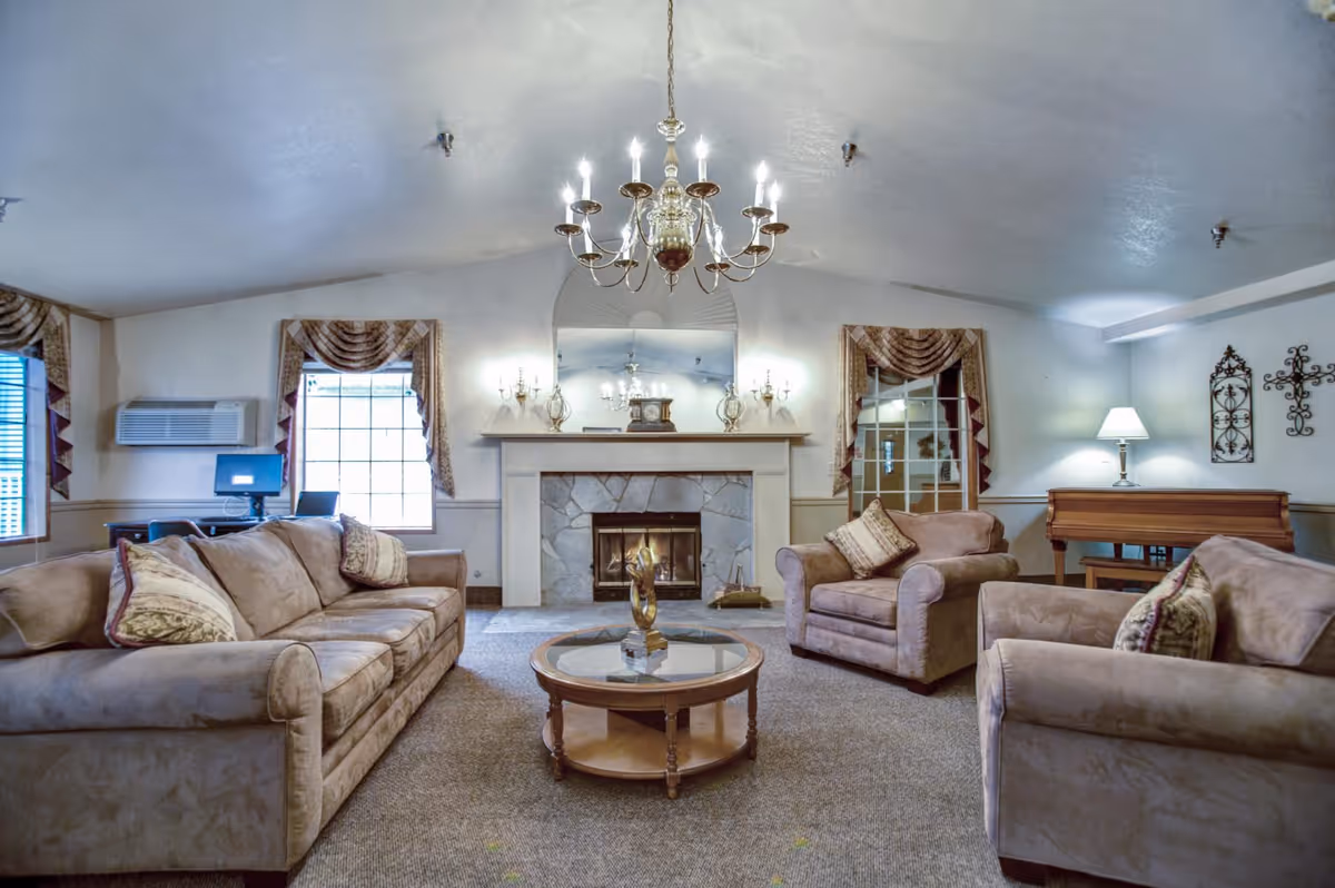 A cozy living room in a senior living facility featuring a beige sofa, two matching armchairs with patterned cushions, a round glass coffee table with a decorative sculpture, a stone fireplace with a mantel clock and candle holders, a chandelier hanging from the ceiling, two windows with draped curtains, a computer desk with a monitor, and a wooden piano with a lamp and wall decorations.