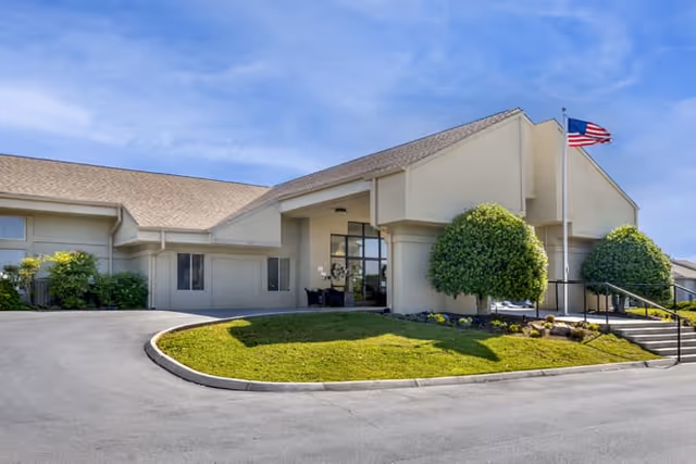 Covered entrance of a single-story senior living facility with manicured landscaping and an American flag beside the driveway.
