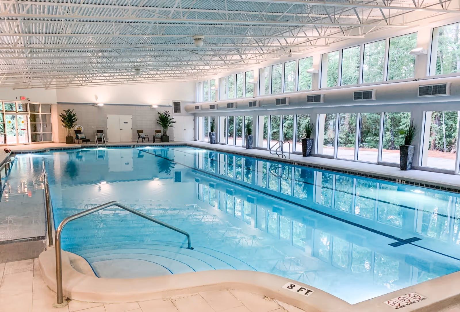 Indoor swimming pool in a bright room with floor-to-ceiling windows, potted plants, and seating along the far wall.