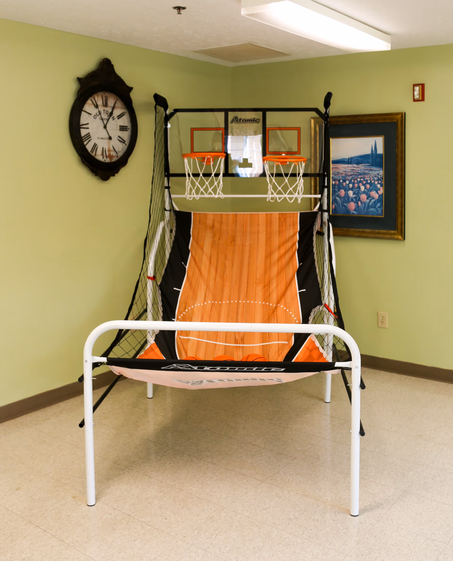 Indoor basketball arcade game with two hoops and a netted frame set up in a room with light green walls, a large wall clock, and a framed painting of a field of flowers.