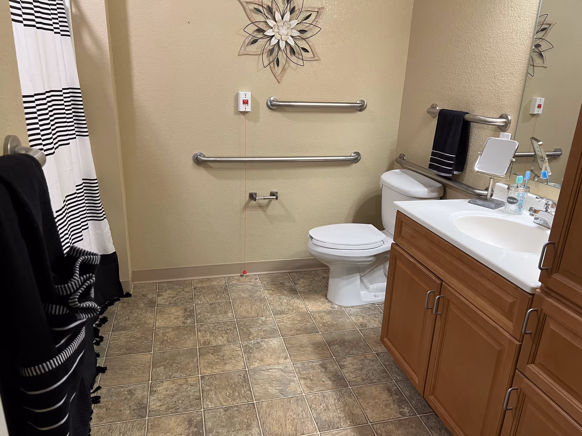 A bathroom with a toilet, a sink with wooden cabinets, a large mirror, and a shower with a black and white striped curtain. There are metal grab bars on the walls near the toilet and a decorative flower wall hanging above the toilet. The floor has brown and beige tiles.