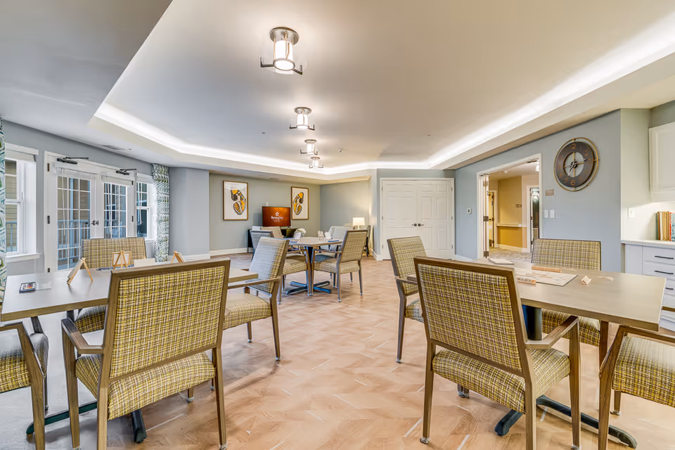 A bright and spacious common area in an assisted living facility with several tables and chairs arranged for social activities or dining. The room features light wood flooring, soft blue walls, modern ceiling lights, and large windows with patterned curtains allowing natural light to enter. There is a clock on the wall and framed artwork, with a doorway leading to another part of the facility.