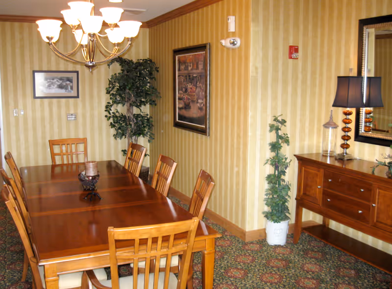 A dining room with a long wooden table surrounded by eight wooden chairs with cushioned seats. A chandelier with multiple lights hangs above the table. The walls are decorated with vertical striped wallpaper, a framed painting, and a framed photograph. There are two potted plants, one in the corner and one near a wooden sideboard with drawers. On the sideboard, there is a lamp with a dark shade and decorative items. A large mirror hangs above the sideboard. The floor is carpeted with a patterned design.