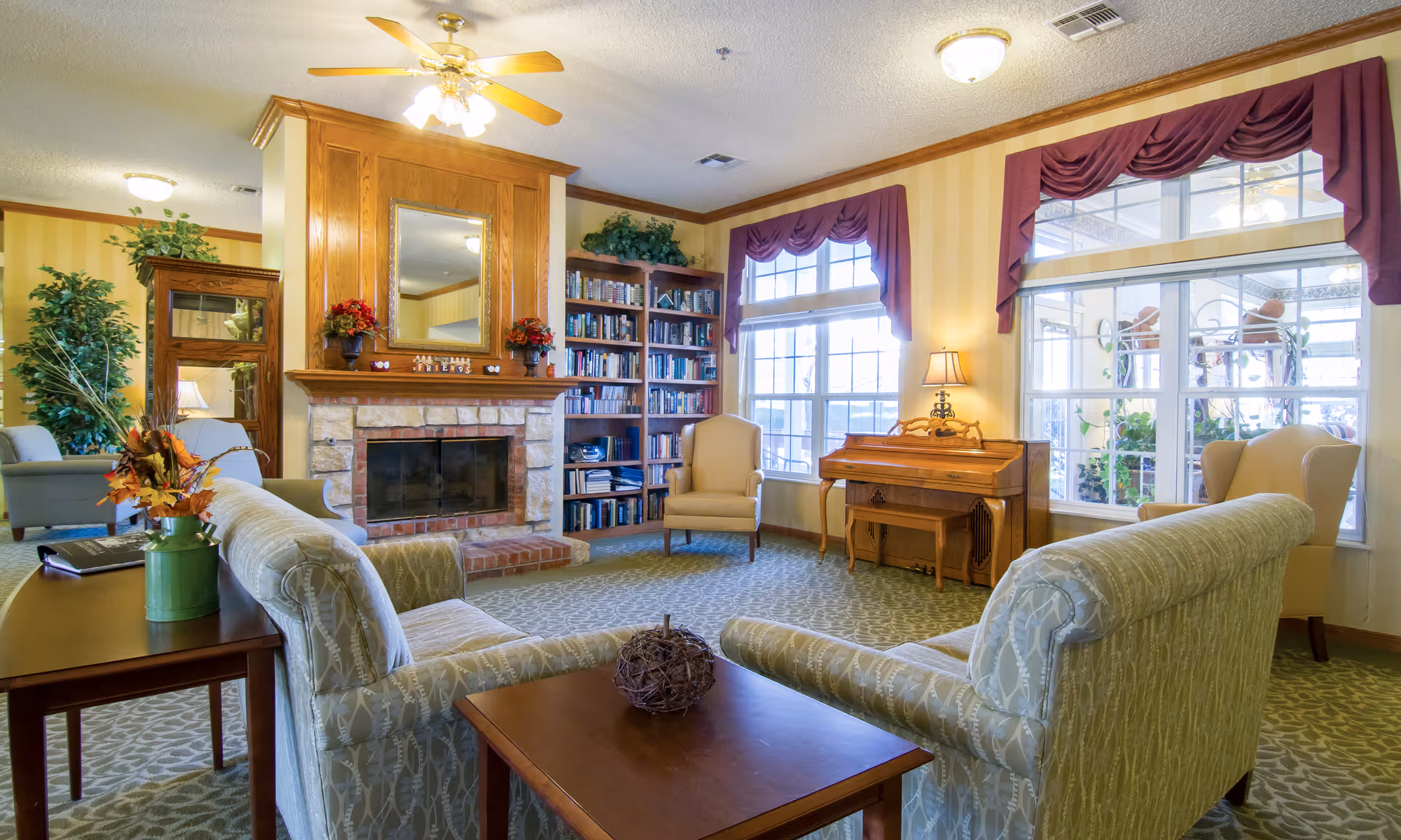 Cozy communal living room with sofas facing a stone fireplace, bookshelves, armchairs, and large windows.