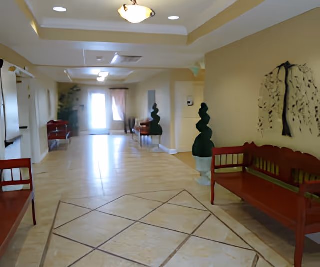 A spacious hallway with beige tiled floors and cream-colored walls. There are red wooden benches along the walls and two topiary plants in white pots. A decorative wall art piece resembling a tree is mounted on the right wall. The hallway is well-lit with ceiling lights and has a door with a window at the far end letting in natural light.