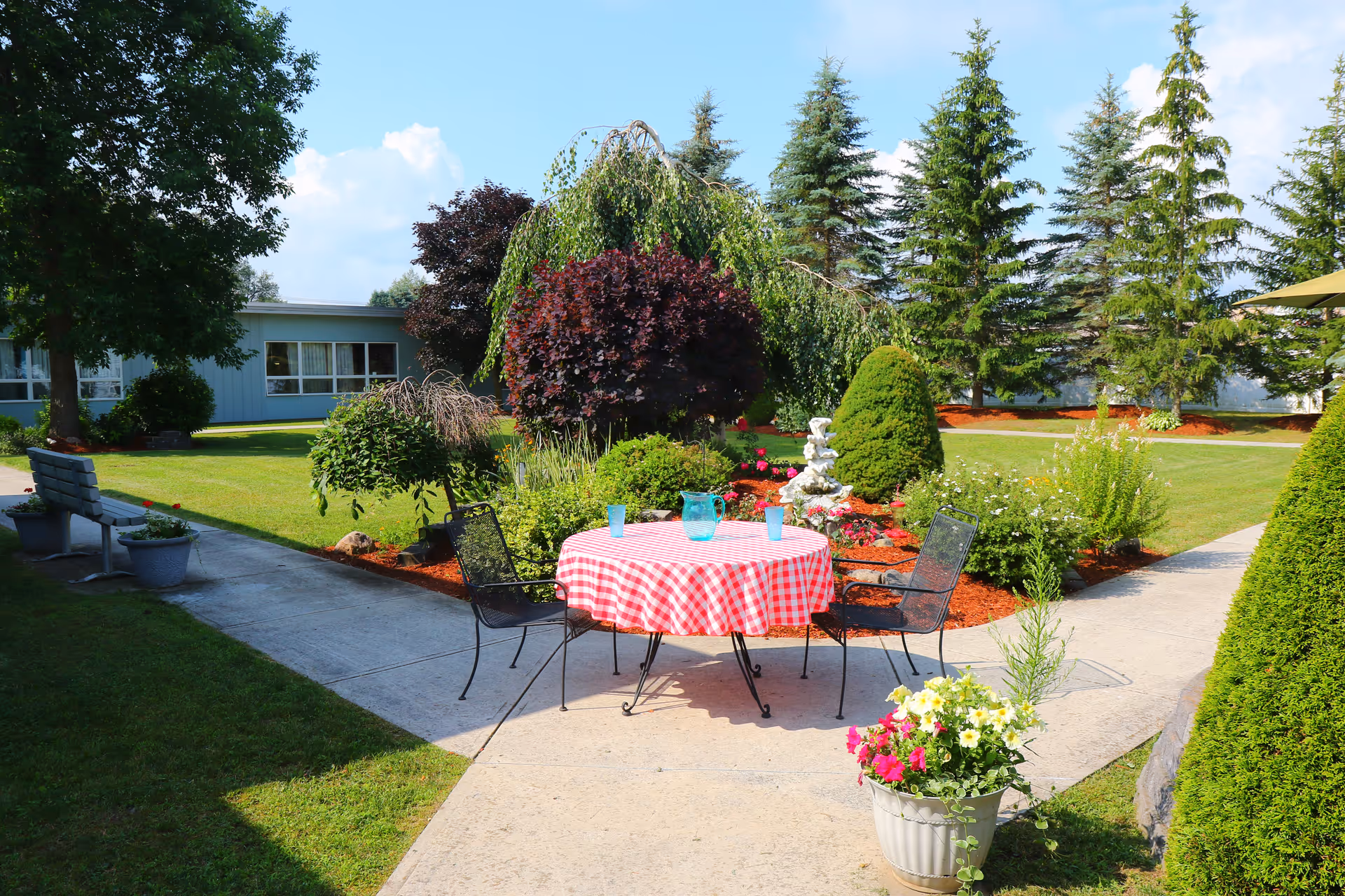 A landscaped outdoor courtyard with a round table covered in a red-and-white checkered cloth, metal chairs, potted flowers, and surrounding trees and shrubs.