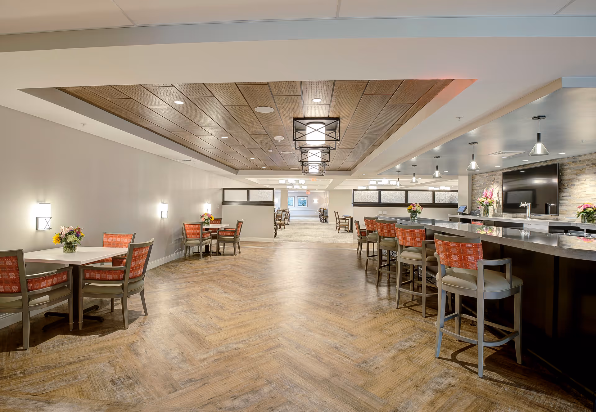 A spacious dining area with wooden flooring and a wooden ceiling panel featuring modern light fixtures. There are several tables with chairs that have red patterned cushions, and a long bar counter with high chairs on the right side. The area is well-lit with wall sconces and pendant lights, and decorated with flower arrangements on the tables and counter.