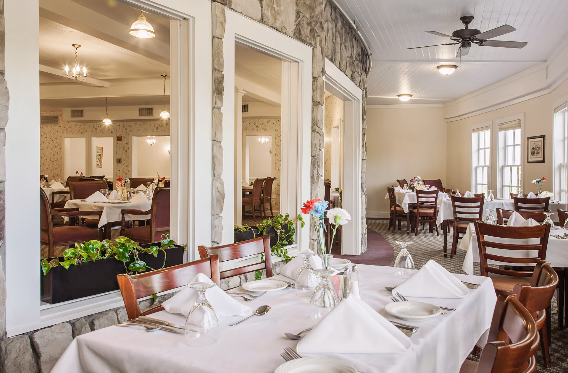 A bright dining room with tables covered in white tablecloths, set with plates, silverware, napkins, and glassware. The room features wooden chairs, floral centerpieces, large windows letting in natural light, and stone-framed interior windows looking into an adjacent dining area.
