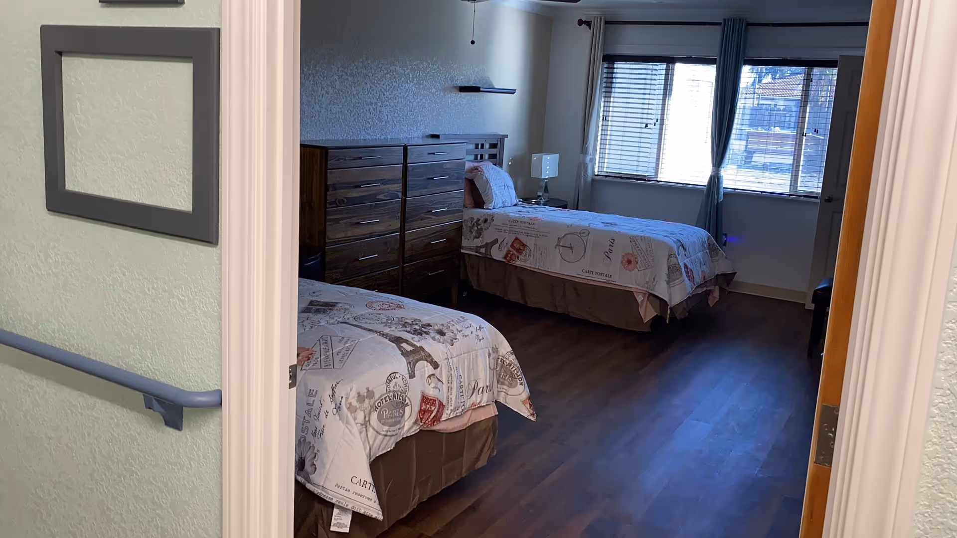 View into a bedroom with two twin beds covered in Paris-themed bedspreads. There is a wooden dresser between the beds and a window with blinds and curtains letting in natural light. The room has wood flooring and light-colored walls.
