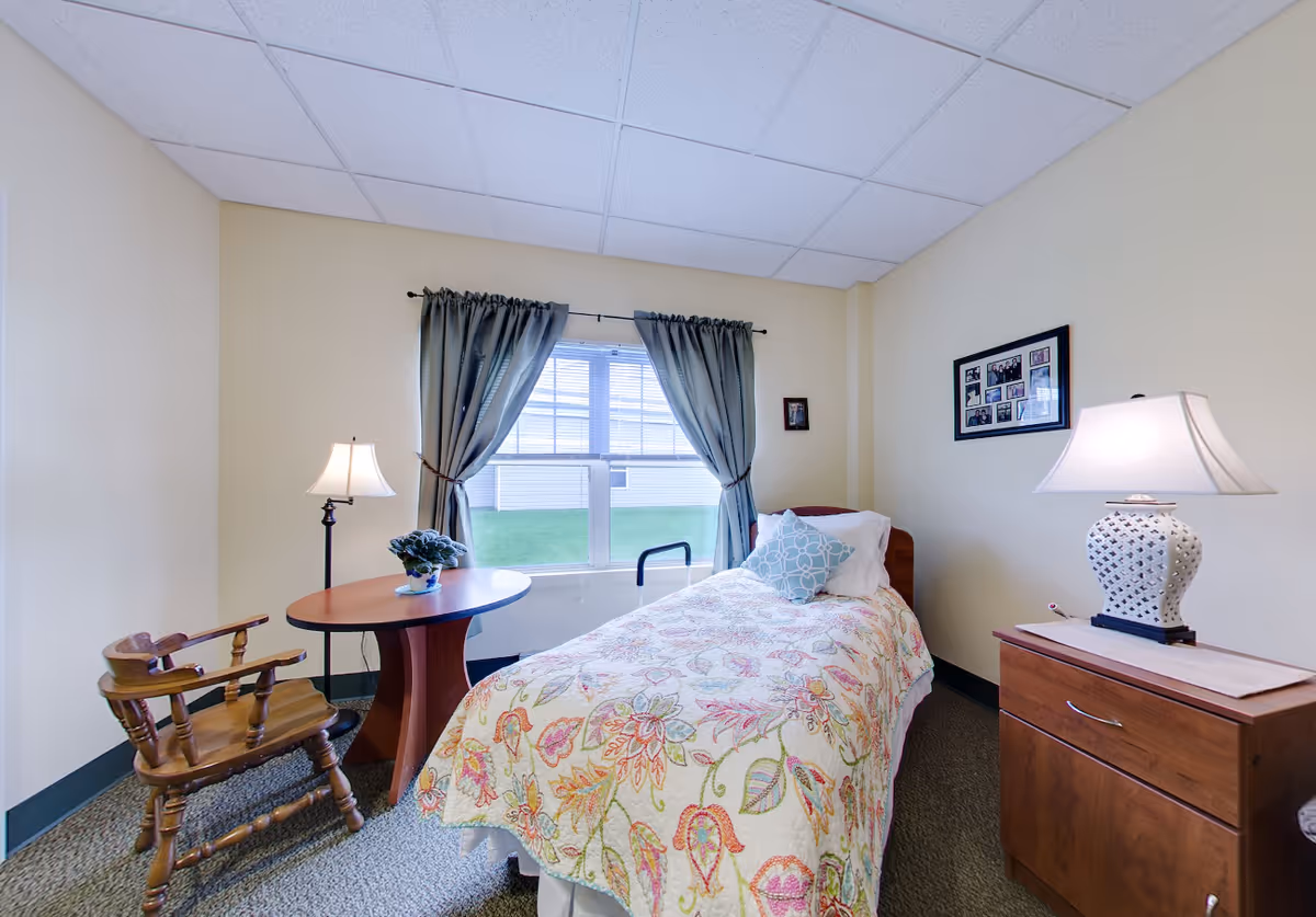 A cozy bedroom in a senior living facility with a single bed covered in a floral quilt, a wooden chair, a round wooden table with a potted plant, two lamps, and a window with blue curtains letting in natural light. There are framed pictures on the walls.