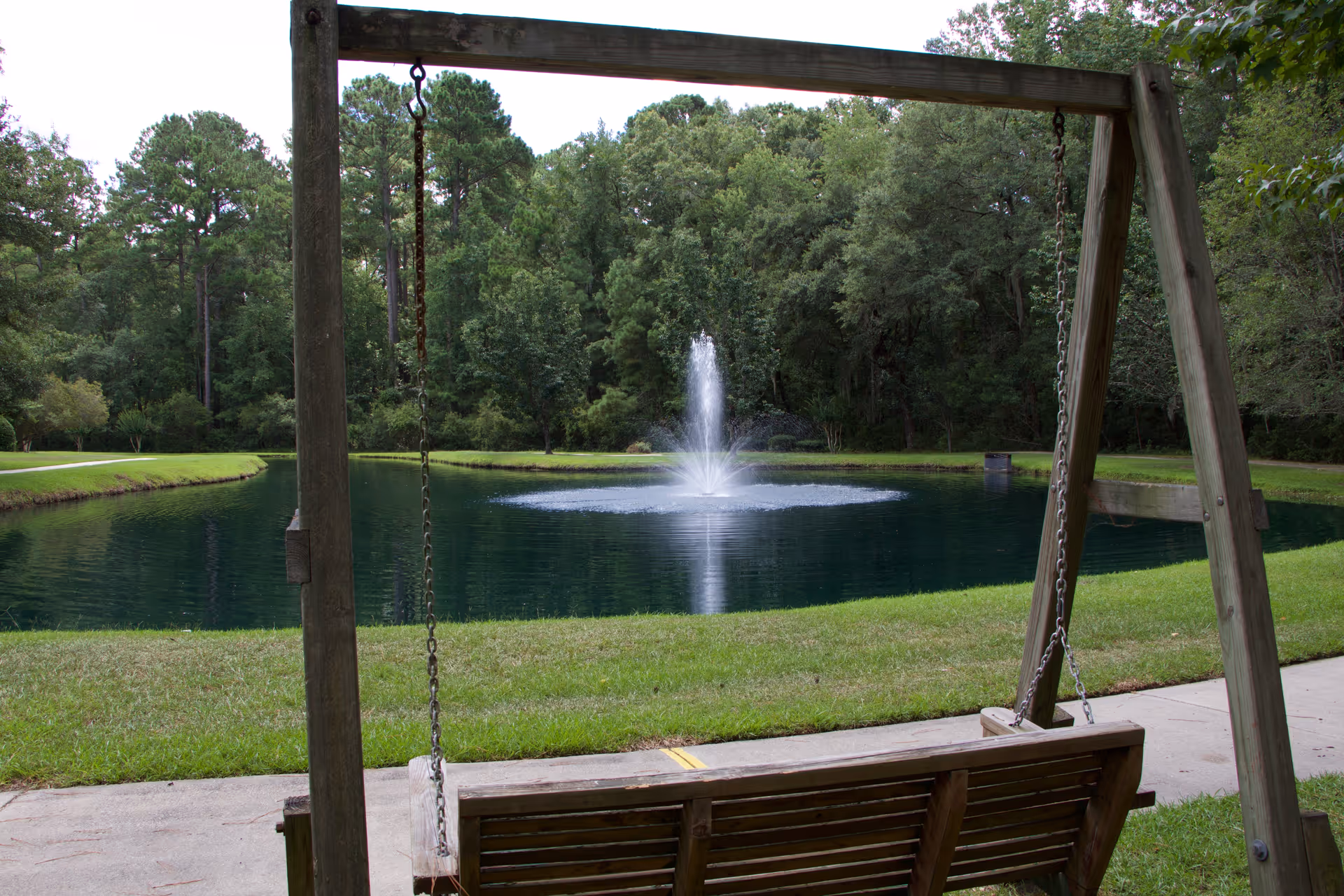 A wooden swing frames a grassy pond with a central fountain and tree-lined background.