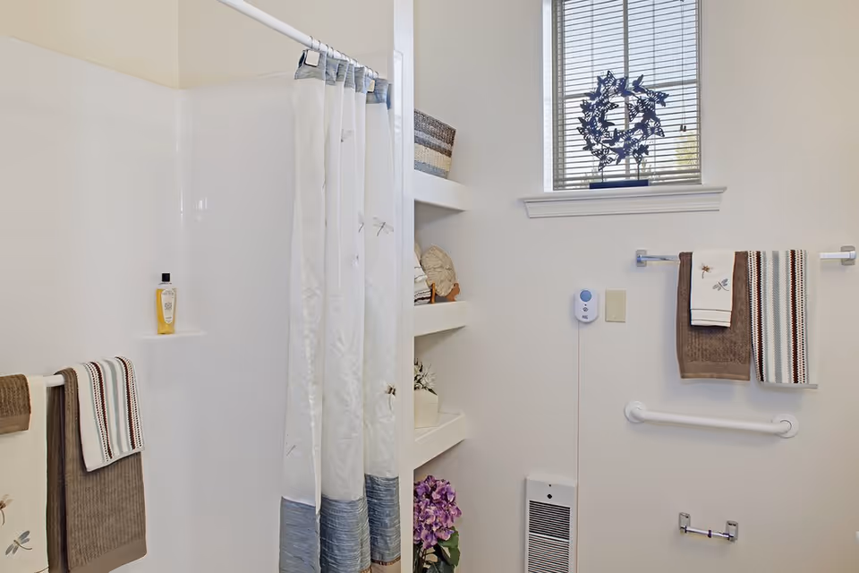 A bright bathroom with a white shower curtain featuring a subtle pattern, a built-in shower shelf holding a bottle of shampoo, and several towels hanging on towel racks. There are built-in shelves with decorative items and a small window with blinds and a decorative wreath. A grab bar and a wall-mounted emergency call button are also visible.