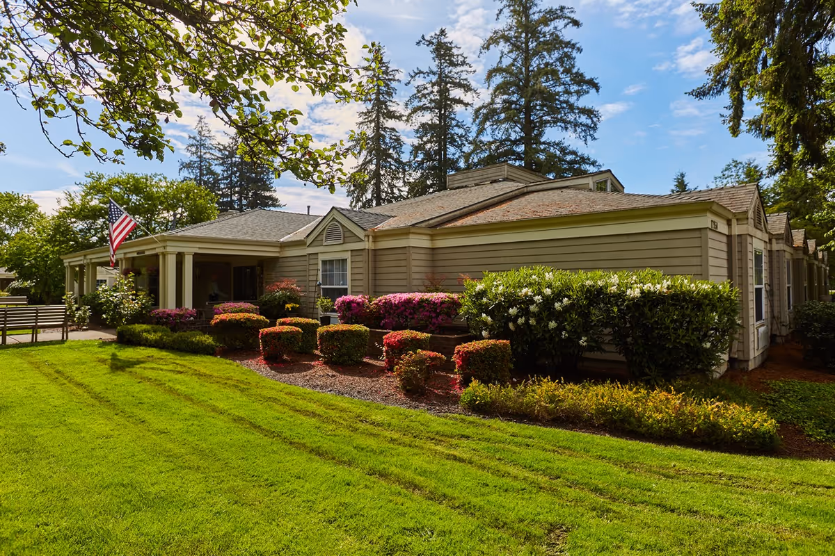 Exterior view of a single-story senior living facility building with beige siding, surrounded by well-maintained green lawn, colorful bushes, and trees under a partly cloudy sky. An American flag is displayed near the entrance.