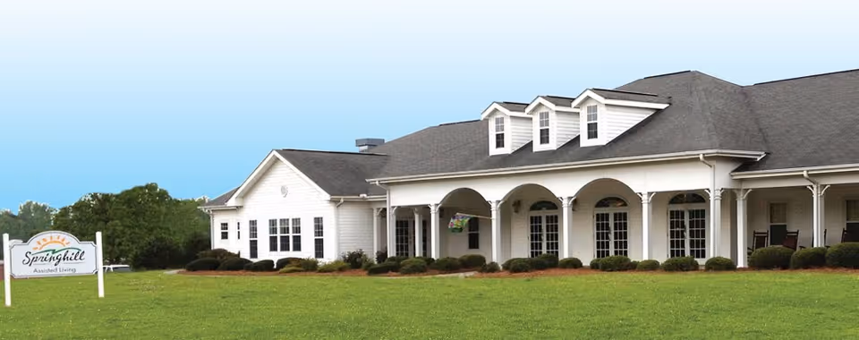 White single-story assisted living building with an arched covered porch, green lawn, and a sign reading "Springhill Assisted Living".