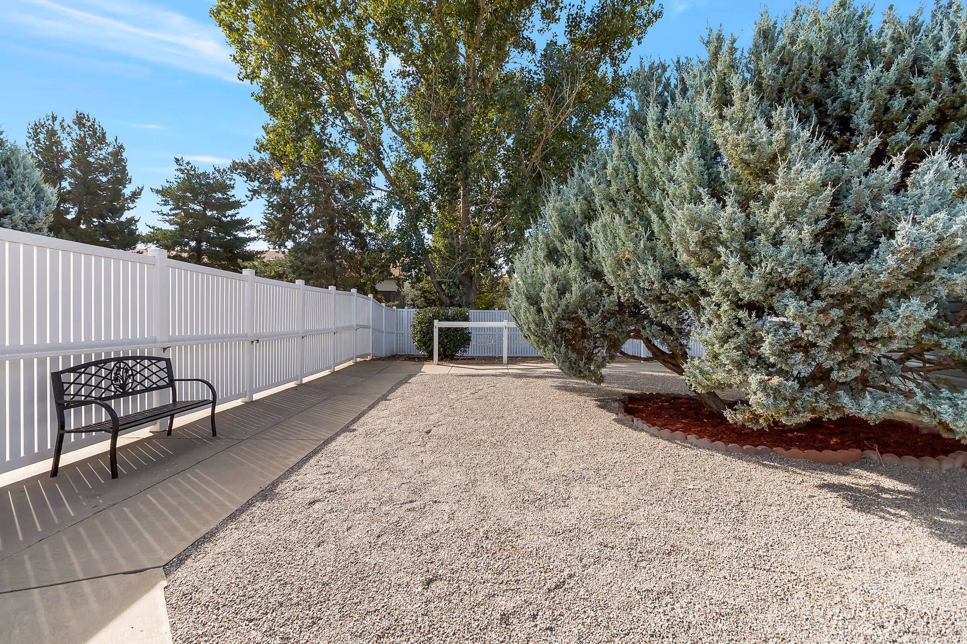 Outdoor area with a gravel ground, a large bush with green foliage on the right, a white fence along the left side, and a black metal bench on a concrete pathway.