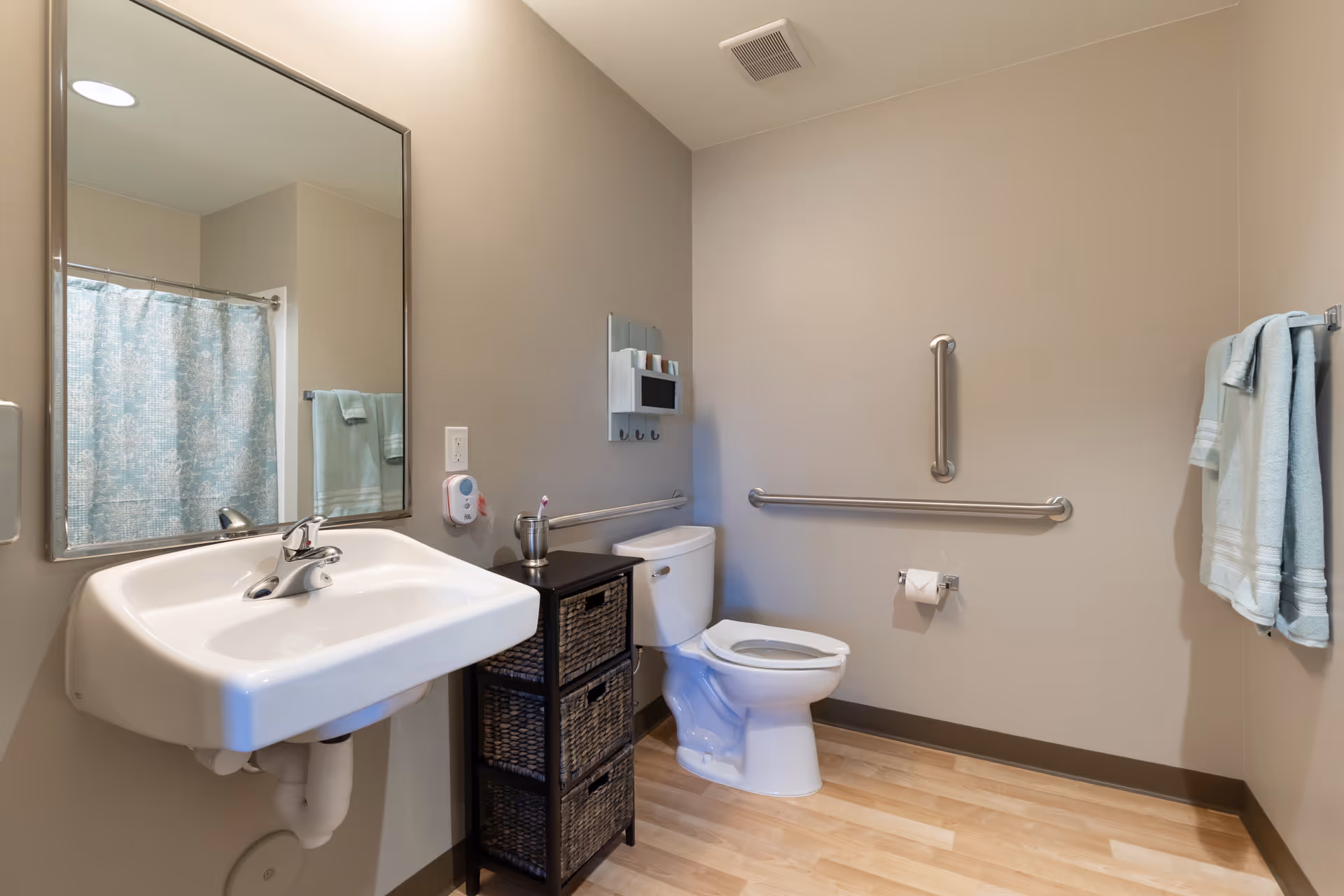 A clean and accessible bathroom with a white wall-mounted sink, a large mirror above it, a toilet with grab bars on the walls for support, a small black storage unit with woven baskets, a towel rack with light blue towels, and a shower curtain visible in the mirror reflection.