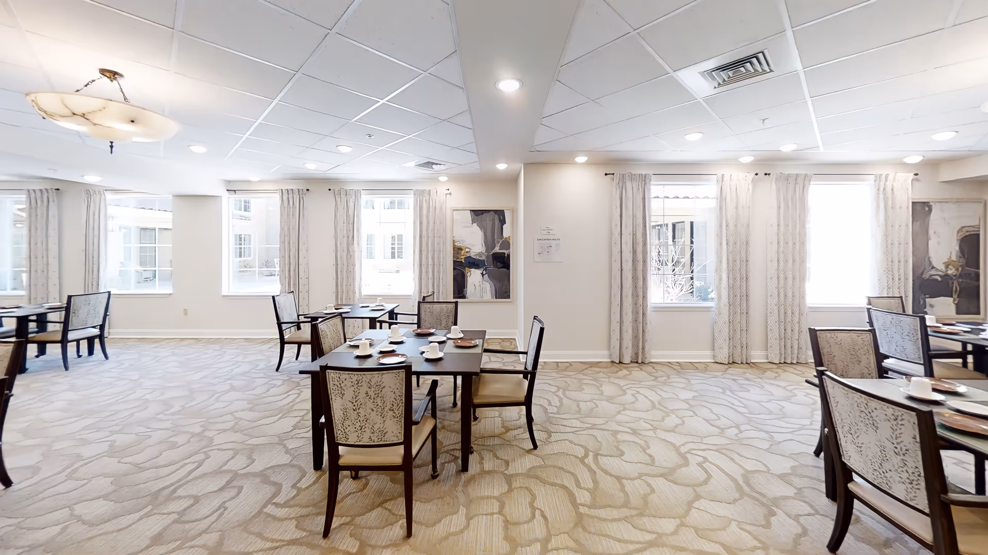 Bright, carpeted dining room with multiple tables and chairs set for meals beneath ceiling lights and large windows.