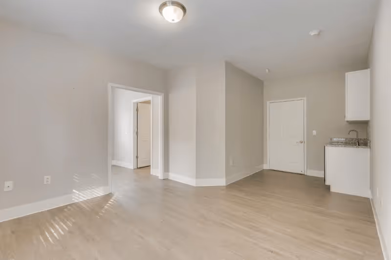 Empty interior room with light wood flooring and beige walls. There is a small kitchenette with a sink and white cabinets on the right side, a closed white door next to it, and an open doorway leading to another room on the left. A ceiling light fixture is visible.