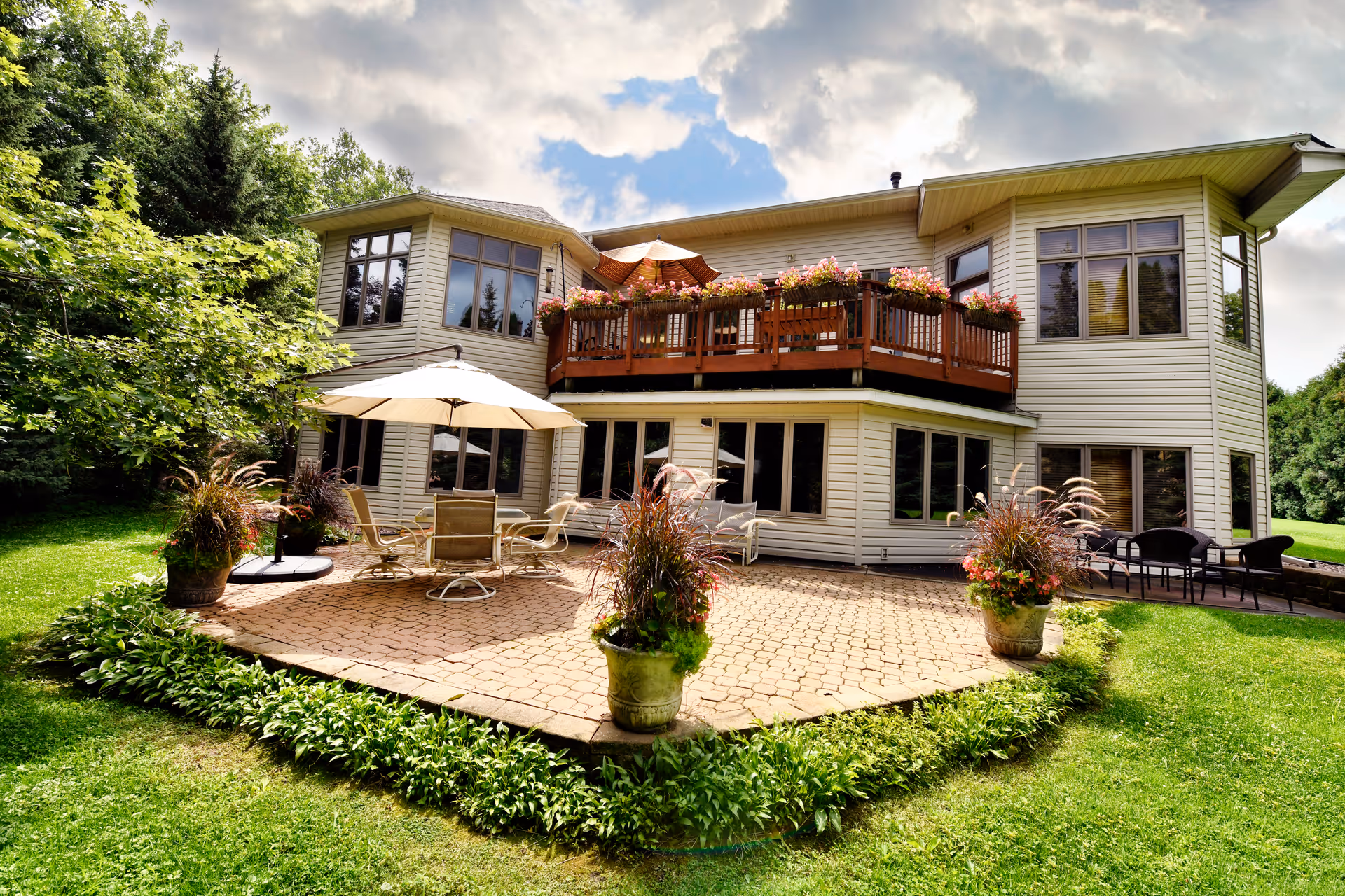 A large two-story house with beige siding surrounded by green grass and trees. The house features a spacious patio area with outdoor furniture including chairs and umbrellas, and a balcony adorned with flower pots. The sky is partly cloudy with patches of blue.