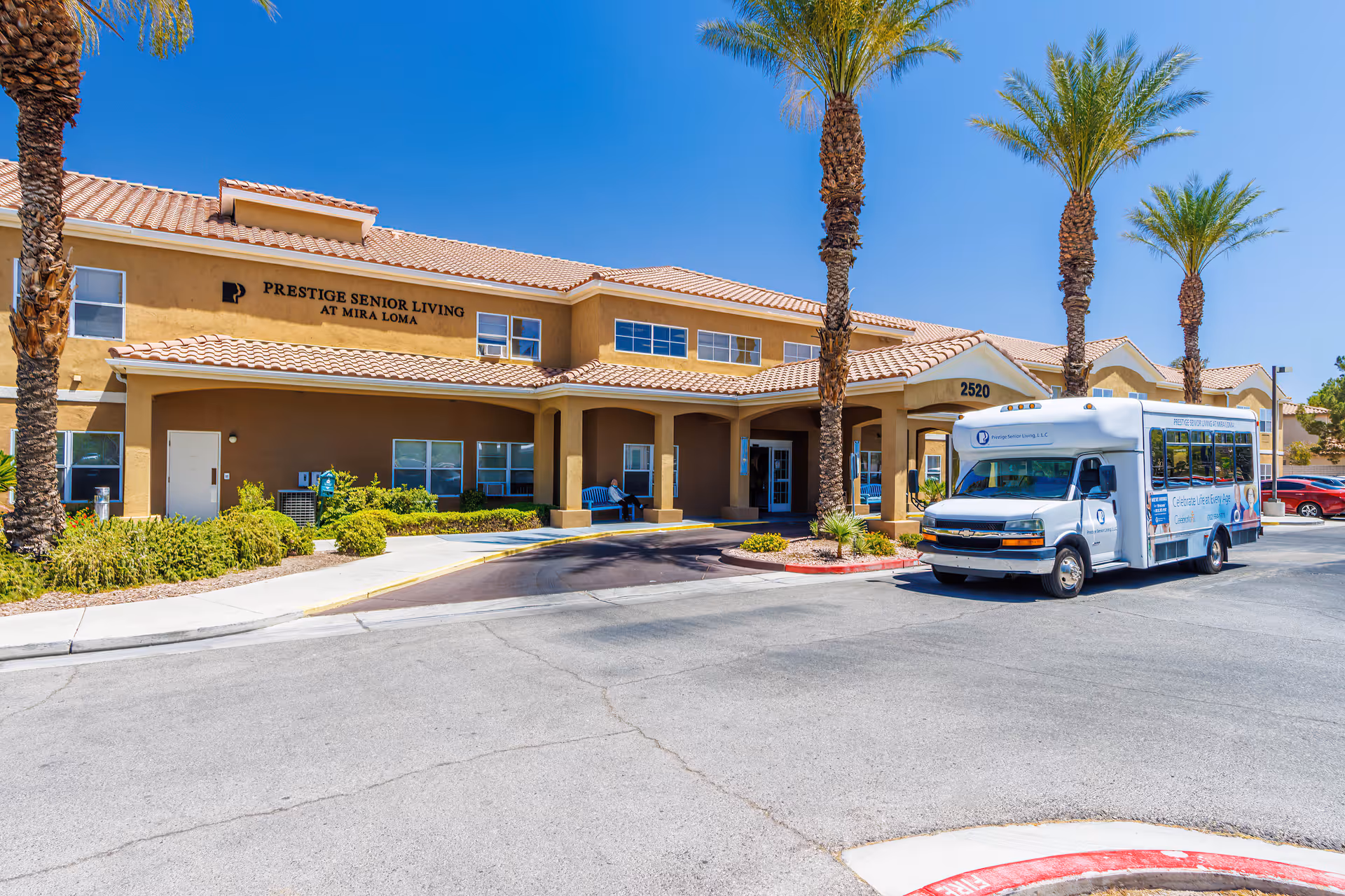 Exterior view of Prestige Senior Living at Mira Loma building with a beige facade and tiled roof under a clear blue sky. There are several tall palm trees in front of the building, a small bus parked near the entrance, and a person sitting on a bench under the covered entryway.