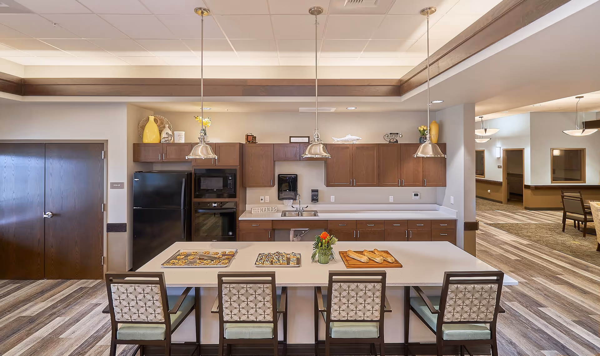 A modern kitchen area in a senior living facility with a large island countertop featuring baked goods and a small flower arrangement. The kitchen has dark wood cabinets, a black refrigerator, a microwave, and an oven. Three pendant lights hang above the island, and there are four chairs with patterned backs and green cushions around the island. The flooring is wood-style, and the room opens into a dining area with tables and chairs.