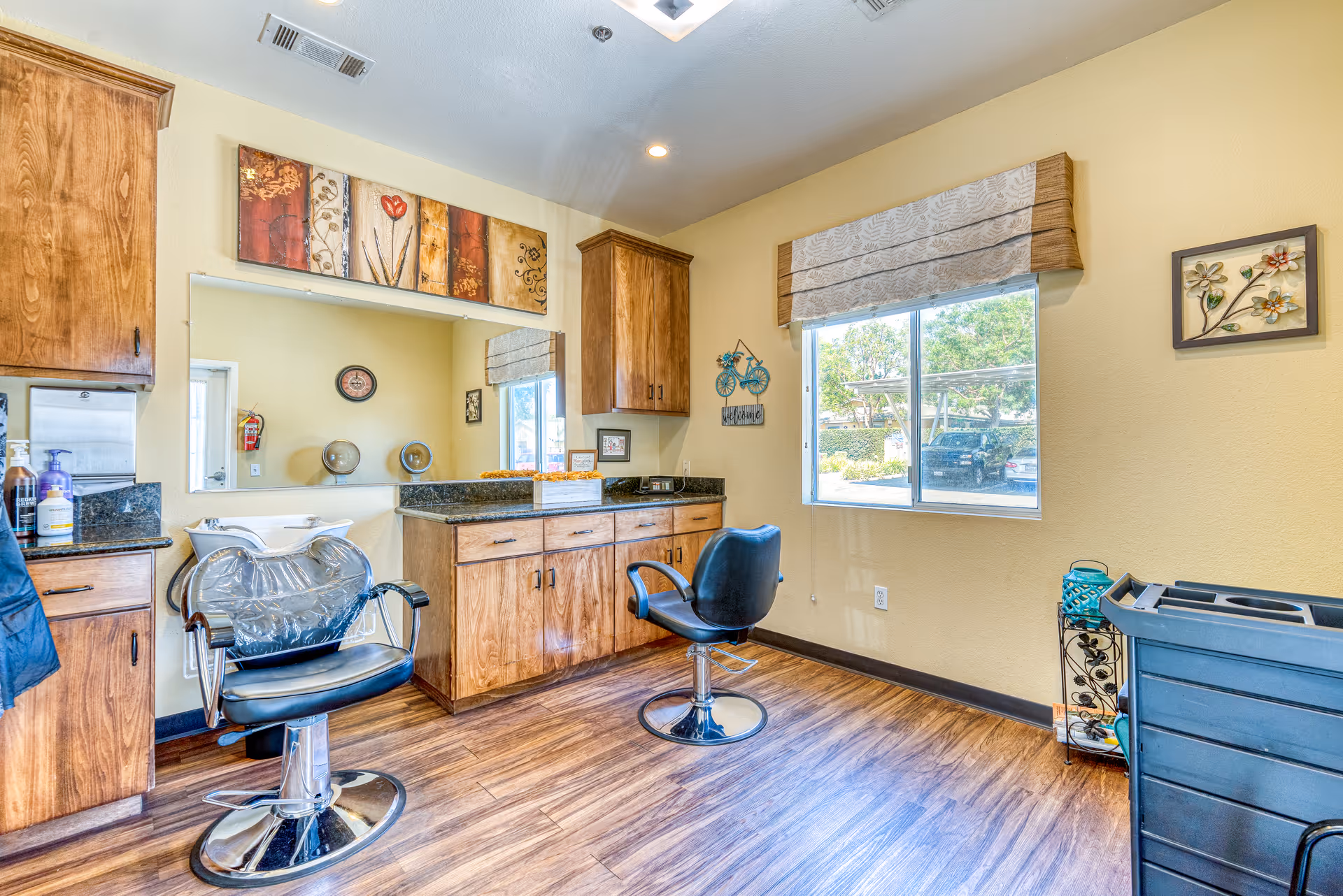 Bright salon-style interior with two styling chairs, wood cabinets, a large mirror, and a window overlooking the parking area.