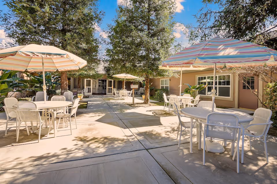 Outdoor patio area at Riverstone Terrace Senior Living with several round tables and white chairs under large striped umbrellas. Trees and plants surround the paved courtyard, and the building with windows and doors is visible in the background under a clear blue sky.