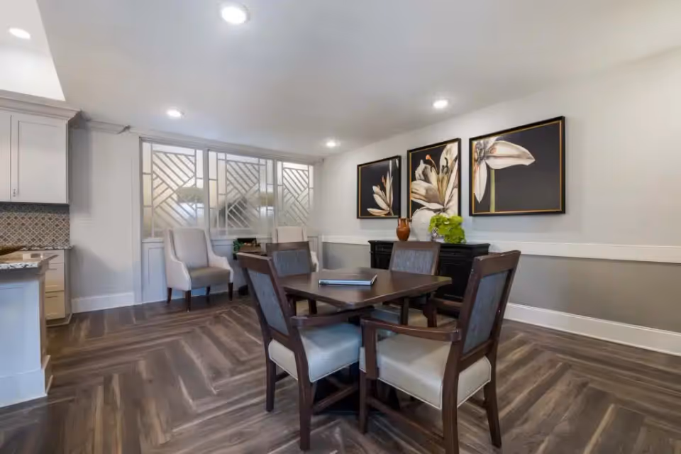 A modern dining area with a square wooden table surrounded by four cushioned chairs. The room features wood-patterned flooring, a sideboard with decorative vases and plants, and three large floral paintings on the wall. Two upholstered armchairs are placed near a decorative glass partition, and the space is well-lit with recessed ceiling lights.
