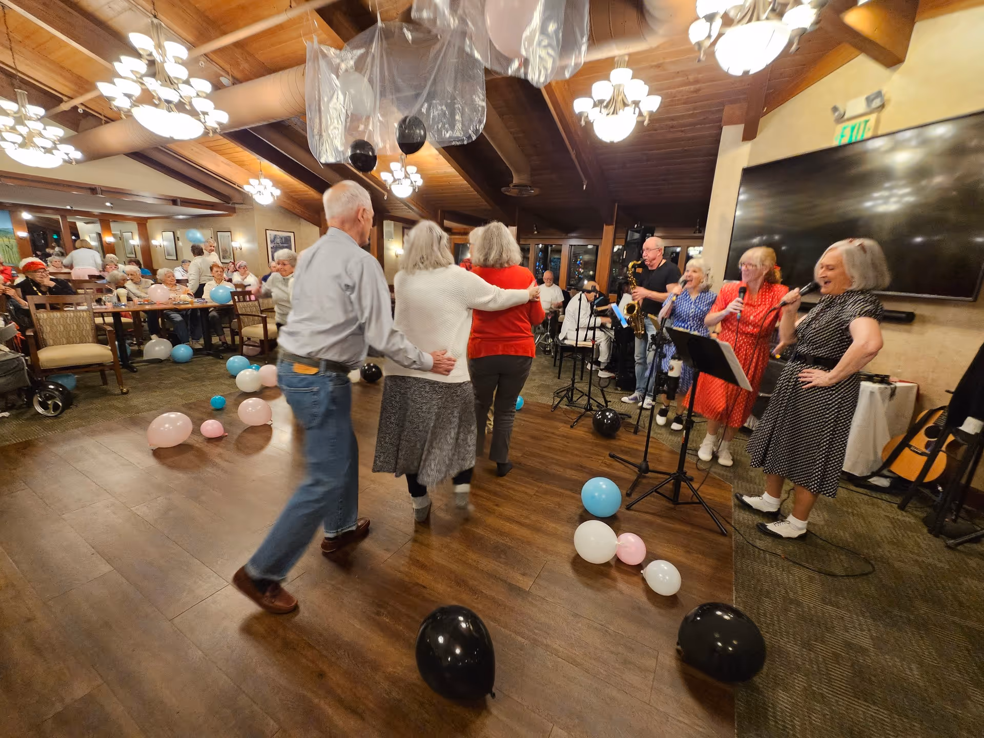 A lively gathering in a senior living facility's common area with elderly residents dancing and enjoying live music. Several people are seated at tables watching the event, while a group of musicians and singers perform near a large TV screen. The room is decorated with balloons in black, white, pink, and blue colors, some floating and some on the floor. The ceiling has wooden beams and multiple light fixtures.