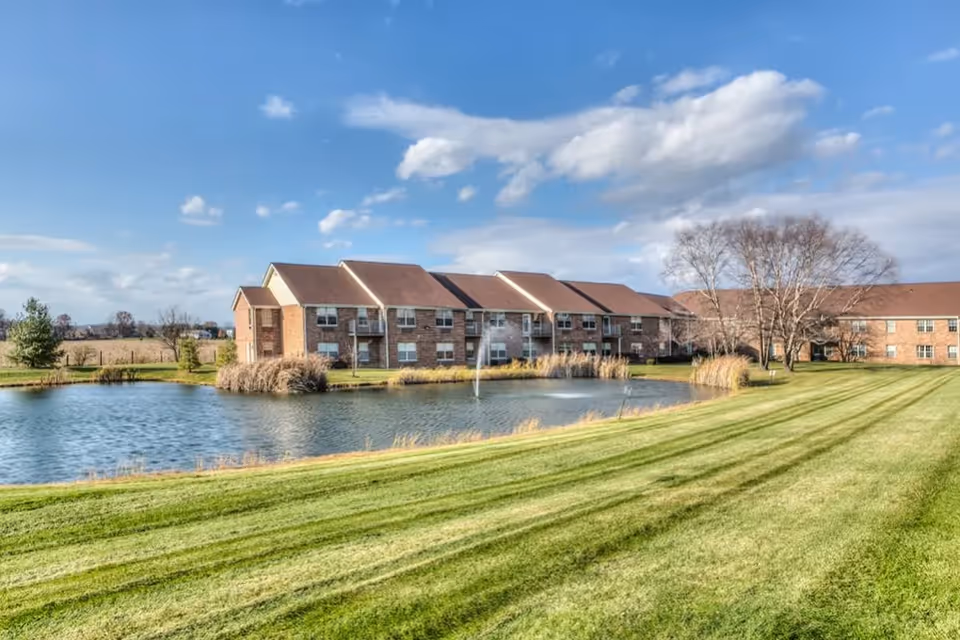 A large brick retirement community building with multiple windows and balconies, situated behind a pond with a water fountain. The foreground features a well-maintained grassy lawn with trees and shrubs under a partly cloudy blue sky.