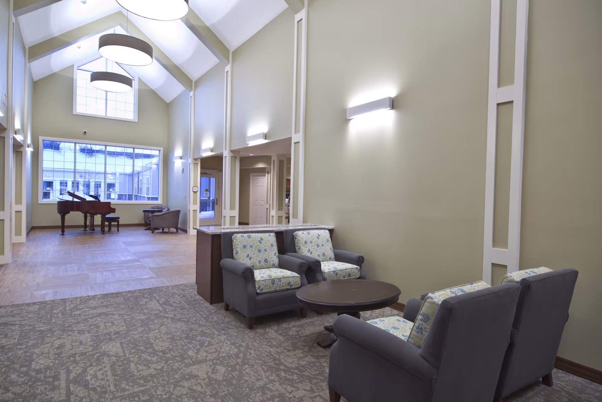 A spacious and well-lit common area with high vaulted ceilings and large windows. The room features a seating area with four cushioned armchairs arranged around a round wooden coffee table. In the background, there is a grand piano near the large windows, and two additional armchairs are placed nearby. The walls are painted a soft beige color, and modern wall sconces provide additional lighting.