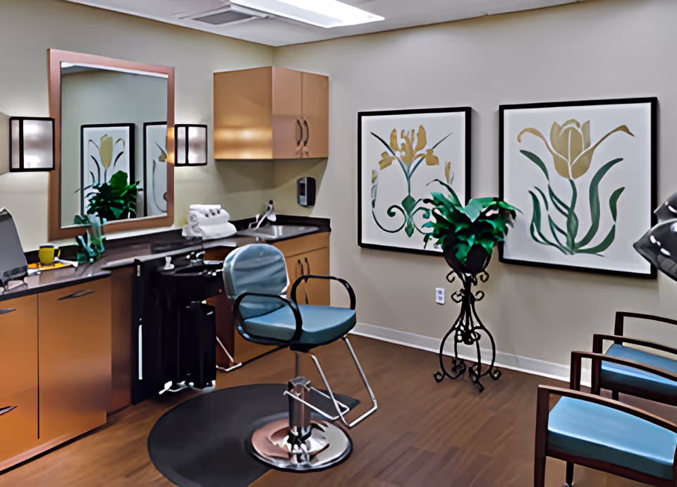 Small salon-style room with a styling chair in front of a mirror, countertop cabinets, framed floral artwork and waiting chairs.