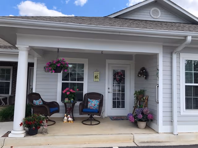 Front porch of a residential building with two brown wicker chairs with blue cushions and floral pillows, a small round table with pink flowers, potted plants on the ground, hanging flower basket, and a door decorated with a wreath. The building exterior is light gray with white trim and a gabled roof.