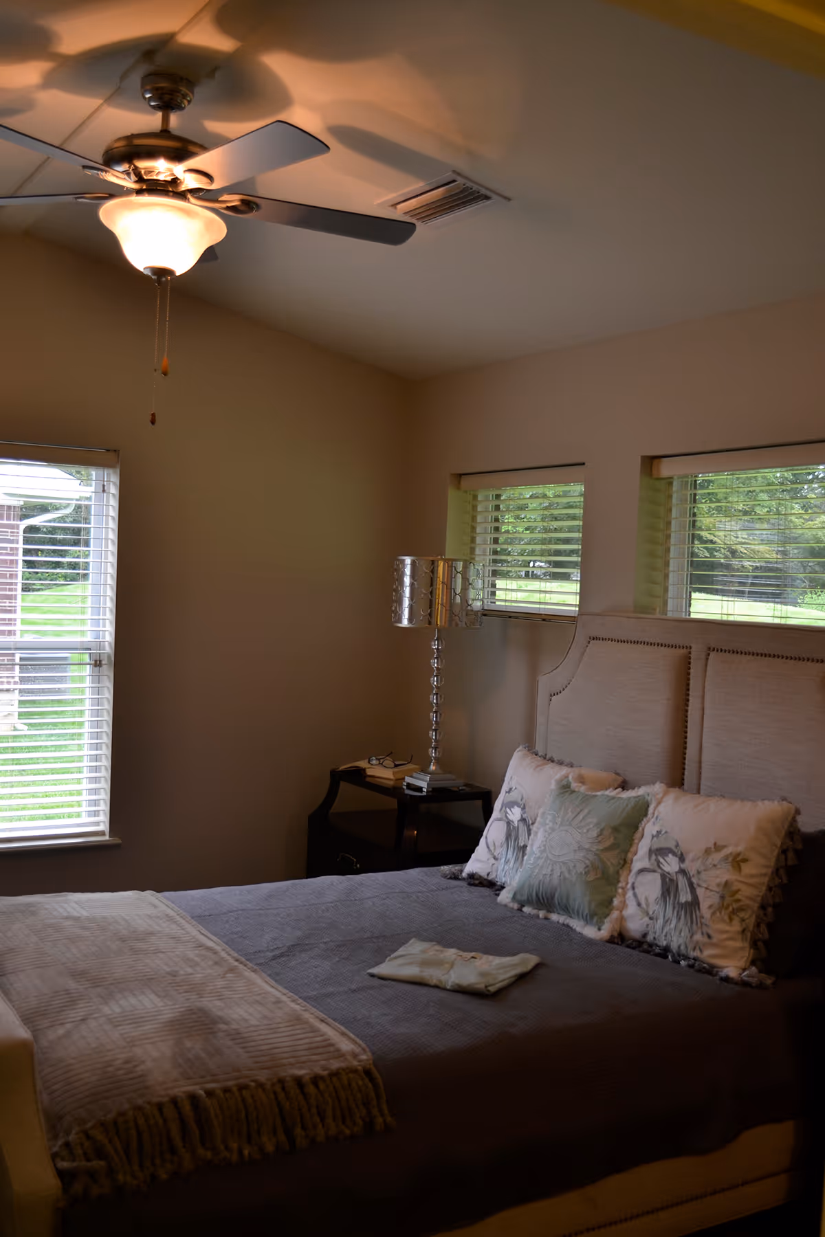 Bedroom with a made bed, decorative pillows, a bedside lamp and nightstand, ceiling fan, and windows overlooking a lawn.